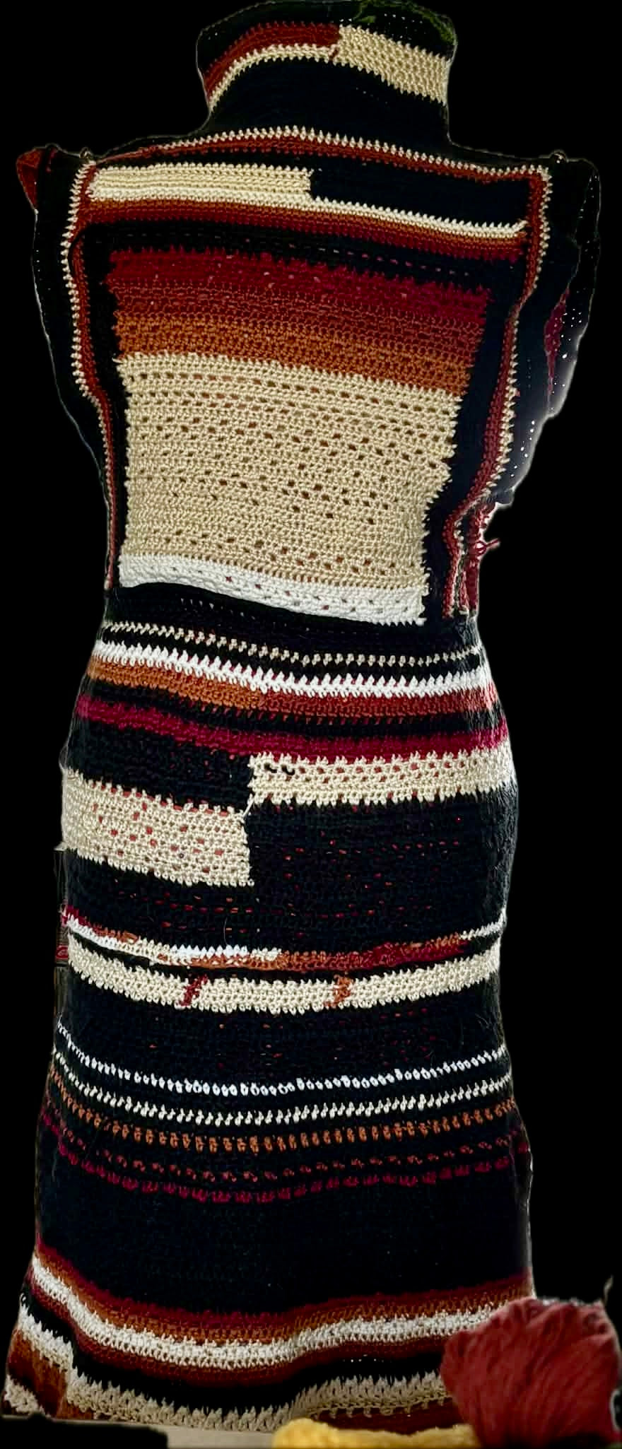 IN PROCESS - Back view of the handmade crocheted Medusa coat on a mannequin, displaying an intricate geometric patchwork design in black, cream, beige, and deep rust red yarn.